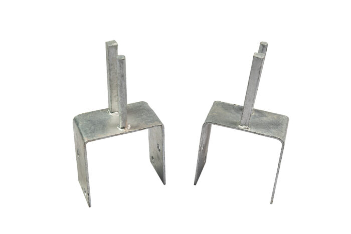A side-by-side comparison of two galvanized steel Jersey barrier saddle brackets on a white background, showing a dual-peg model on the left and a single-peg model on the right.