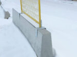 A close-up view showing the bottom frame of a yellow temporary fence panel slotted over the vertical pegs of galvanized saddle brackets attached to a concrete Jersey barrier.