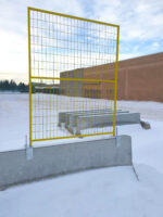 A yellow temporary fence panel securely mounted on top of a concrete Jersey barrier using heavy-duty galvanized steel saddle brackets in a snowy outdoor setting.