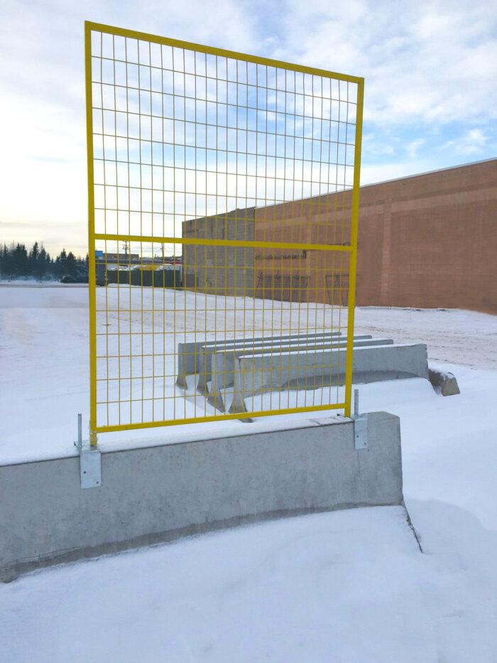 A yellow temporary fence panel securely mounted on top of a concrete Jersey barrier using heavy-duty galvanized steel saddle brackets in a snowy outdoor setting.