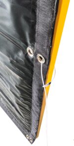 A close-up view of a heavy-duty black acoustic noise barrier installed on a yellow temporary fence, highlighting the reinforced stitched edge and metal grommet used for secure wire tie attachment.